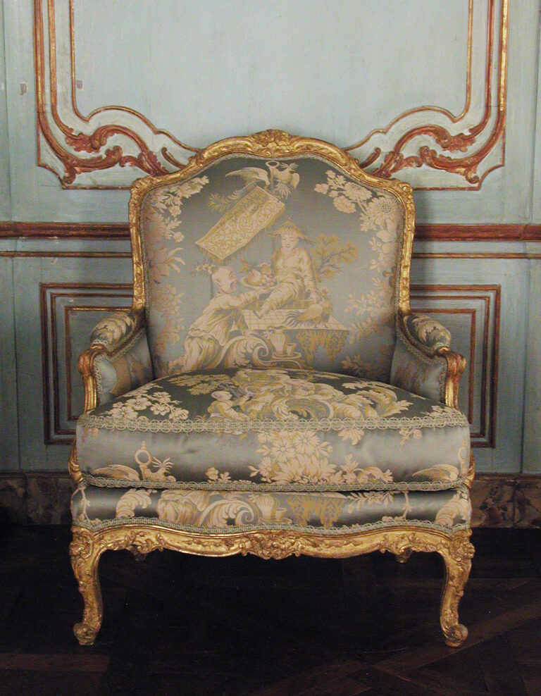 Gilded beechwood bergère with closed, upholstered sides and a loose seat cushion, covered in pale silk with a Chinese-style scene, in front of painted wall panelling