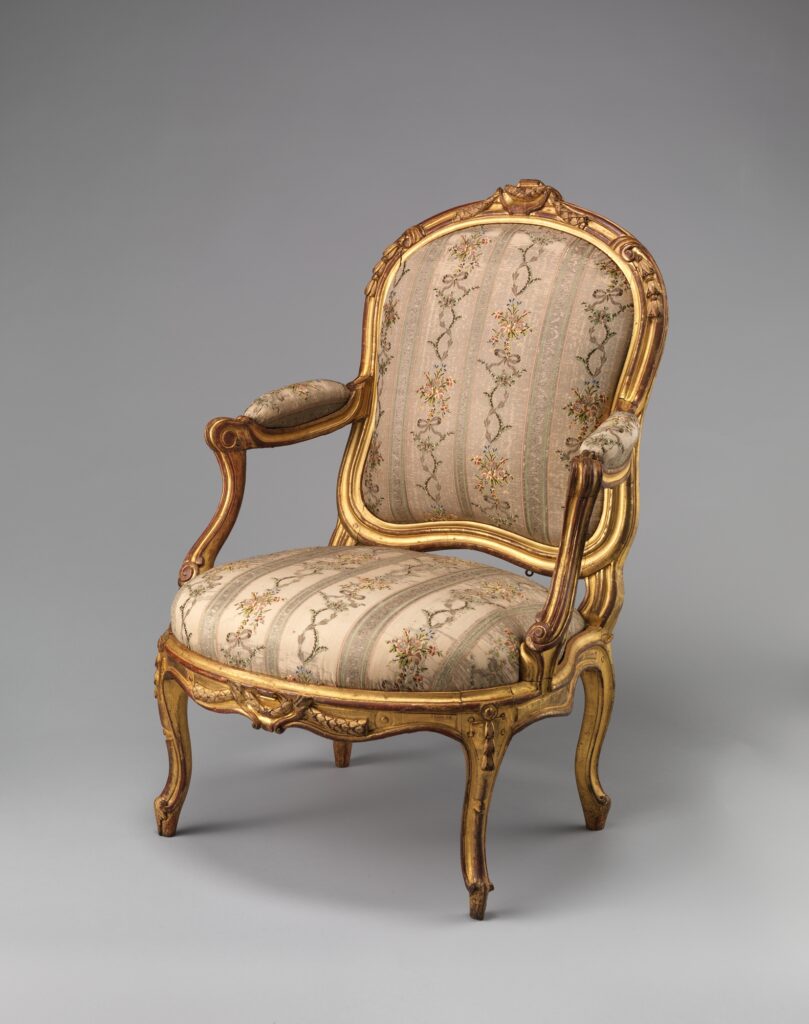 Bergere chair  Gilded walnut fauteuil with open arms, padded arm rests and an upholstered back in a carved gilt frame, seen at an angle against a grey ground