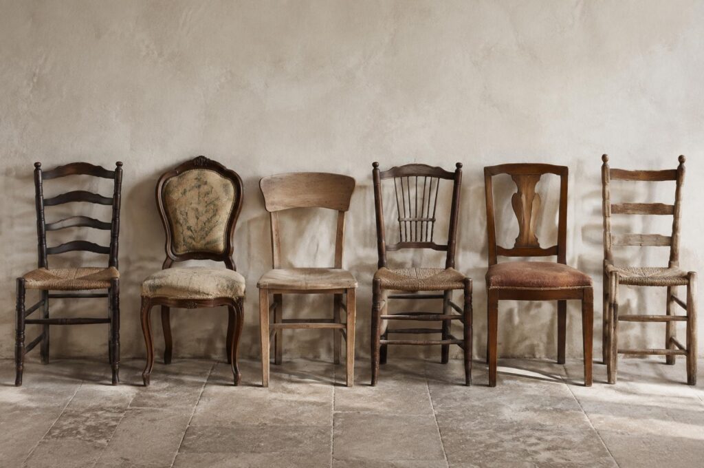 Six mismatched old chairs against a plaster wall, illustrating choices when restoring old chairs.