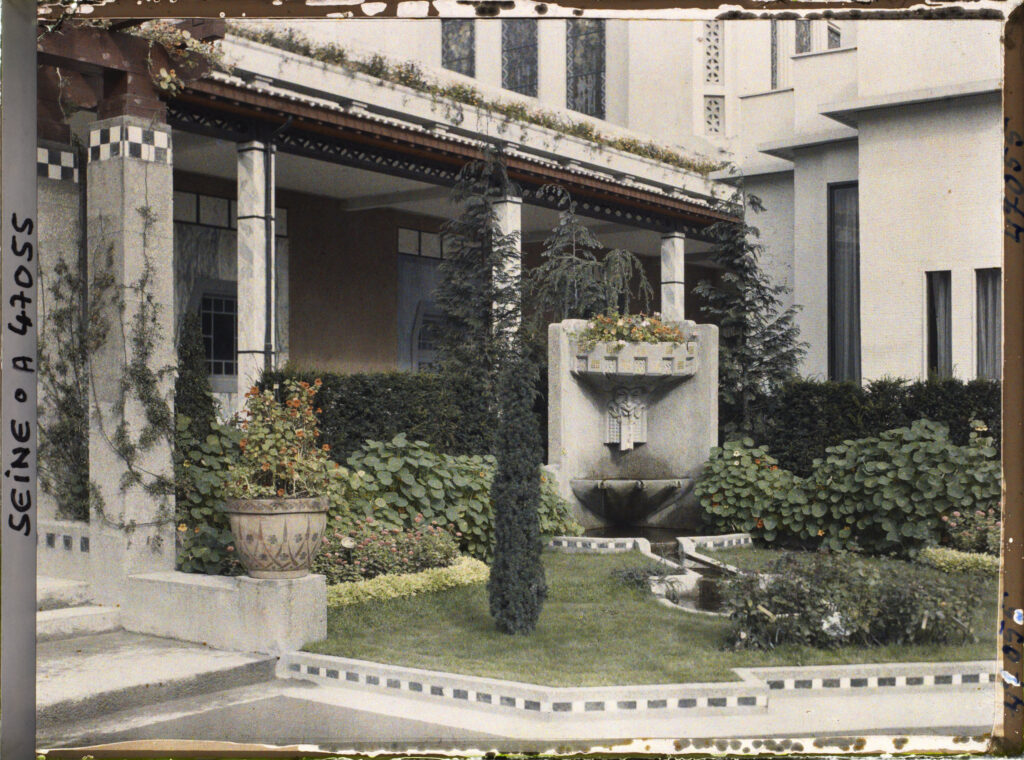 Garden beside the Ruhlmann pavilion in 1925: a loggia with pale columns, a lawn edged with a band of small tiles, clipped hedges and conifers, flowers in pots and a wall fountain, in front of a pale stepped building