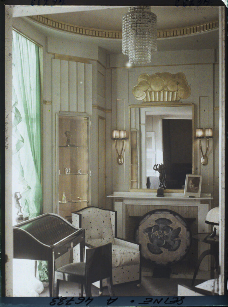 French Art Deco furniture and decoration in Ruhlmann's small salon, 1925 colour plate: pale panelled walls edged in gilt, a tiered crystal chandelier, a gilded relief above a mirror, a dark desk with a slatted lid, a cream armchair and a round painted panel of birds before the fireplace