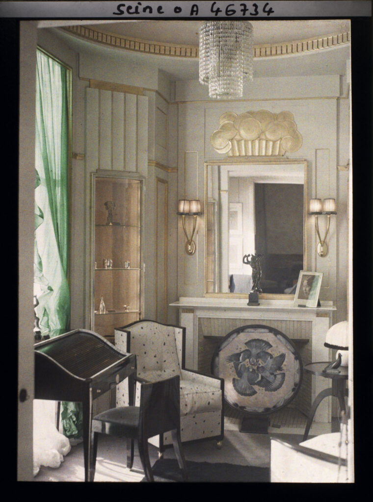 Ruhlmann's small salon in 1925, in a cooler colour plate: pale panelled walls, a tiered crystal chandelier, a gilded relief above a mirror, a dark desk with a slatted lid, a cream armchair and a round painted panel of birds before the fireplace