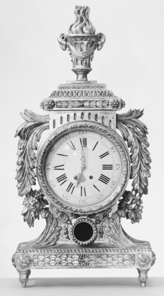 Black-and-white photograph of a tall mantel clock with an urn finial on a carved, gilded wood case with leaf scrolls around a round dial