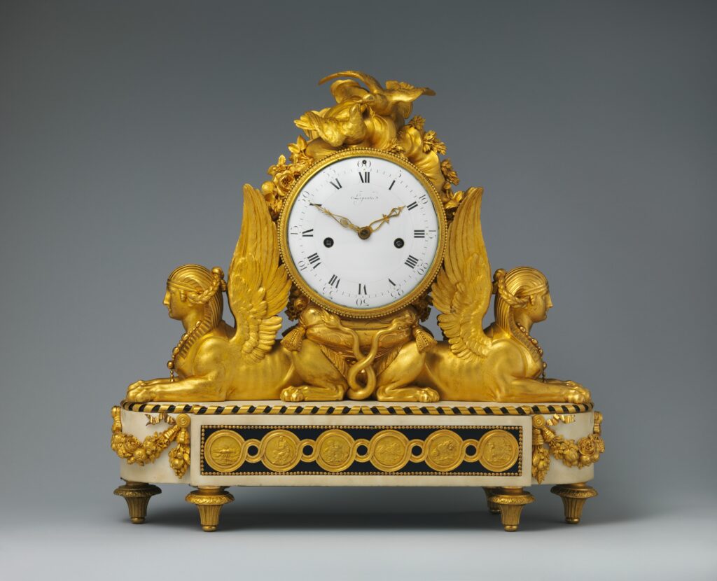 Mantel clock with a white enamel dial between two winged sphinxes in gilt bronze, on a white marble base with a gilt-bronze frieze of medallions, against a grey ground