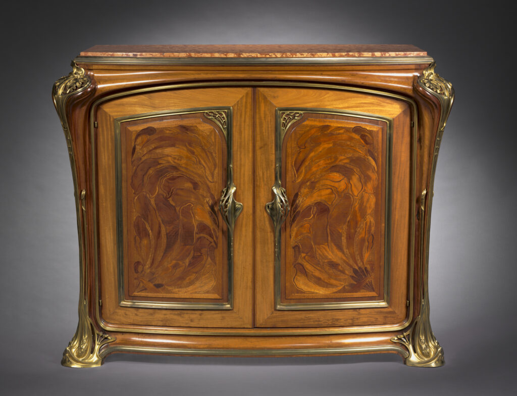 Low two-door cabinet of polished wood with gently curving sides, swirling reddish marquetry on both doors and gilt-metal mounts at the corners, photographed in a grey studio