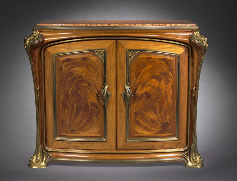 Low two-door cabinet of polished wood with gently curving sides, swirling reddish marquetry on both doors and gilt-metal mounts at the corners, photographed in a grey studio
