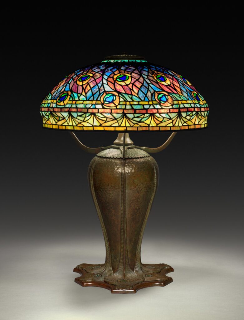 Tiffany Lamp Domed leaded-glass shade patterned with peacock-feather eyes in blue, green and orange, on a dark bronze base with a swelling body and leaf-shaped feet