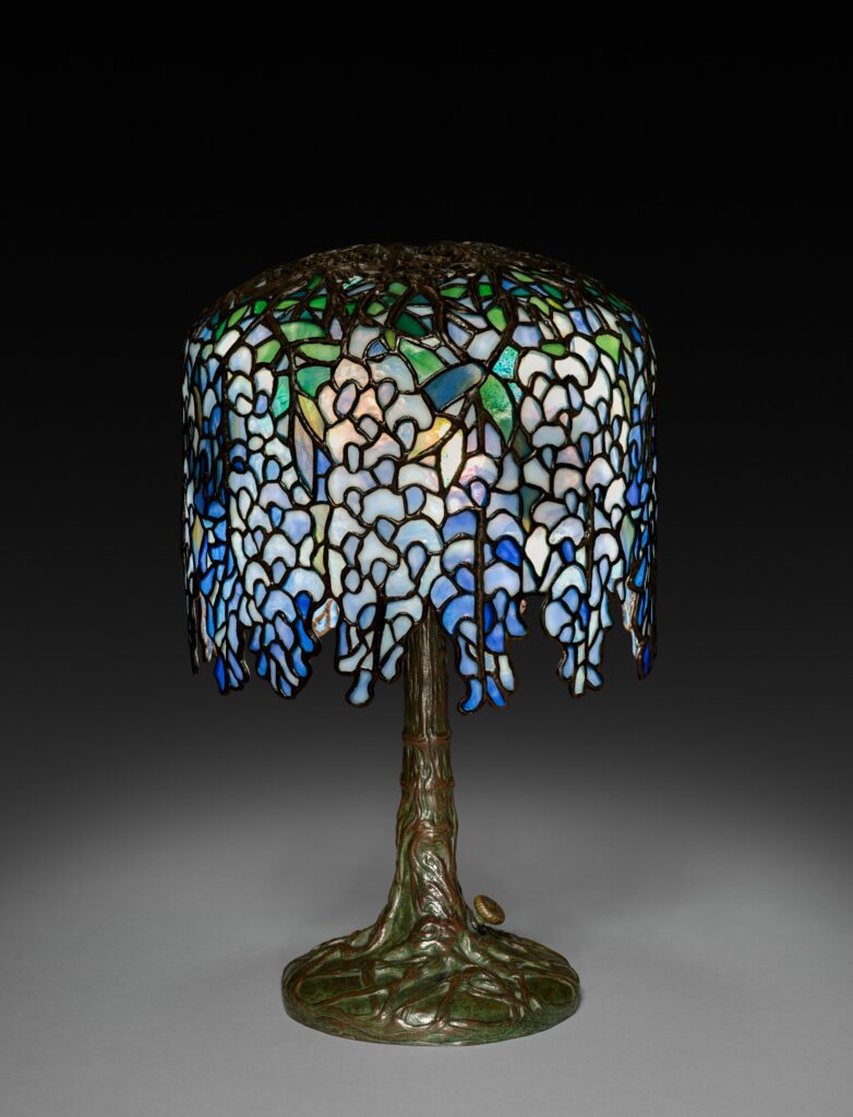 Table lamp with a domed shade of blue and white glass pieces hanging like wisteria blossoms, on a bronze base shaped like a tree trunk with roots spreading at the foot