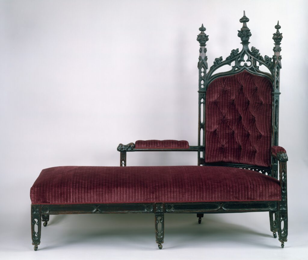 Long low couch in buttoned burgundy upholstery with a tall pointed-arch back at one end, carved with pinnacles and pierced tracery, on a dark green frame with casters