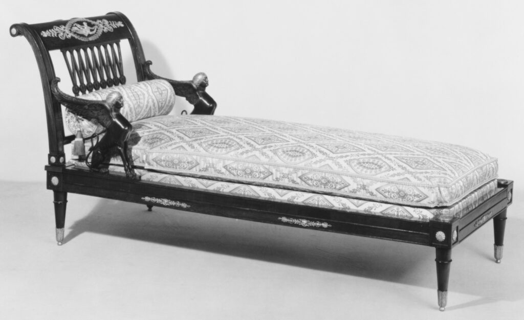 Black and white photograph of a mahogany daybed with a scrolled end, sphinx-shaped arm supports, a pale patterned cushion and slim tapering legs