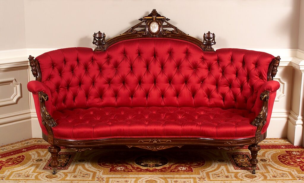Red satin sofa with a deeply buttoned back and seat, a carved wooden crest with a central oval medallion and small finials, and short turned legs, on a patterned carpet
