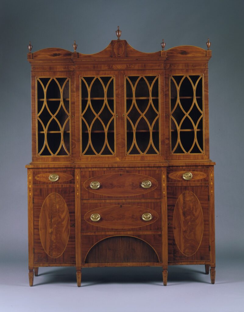Wide American mahogany secretary of 1800-1810 with four glazed upper doors set with curved lattice glazing bars under a shaped crest with finials, above a lower case of drawers and cupboards with oval veneer panels