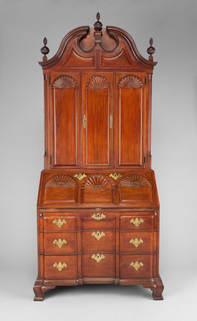 Tall American mahogany desk and bookcase of 1760-90 with a scrolled broken-pediment top and three finials, fan-carved shell designs on the doors and the writing section above three tiers of drawers with brass handles
