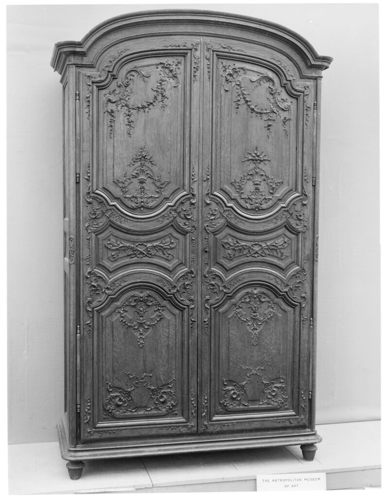 Black-and-white photograph of an antique French armoire with two carved oak doors in shaped panels with floral swags, a curved cornice and short feet