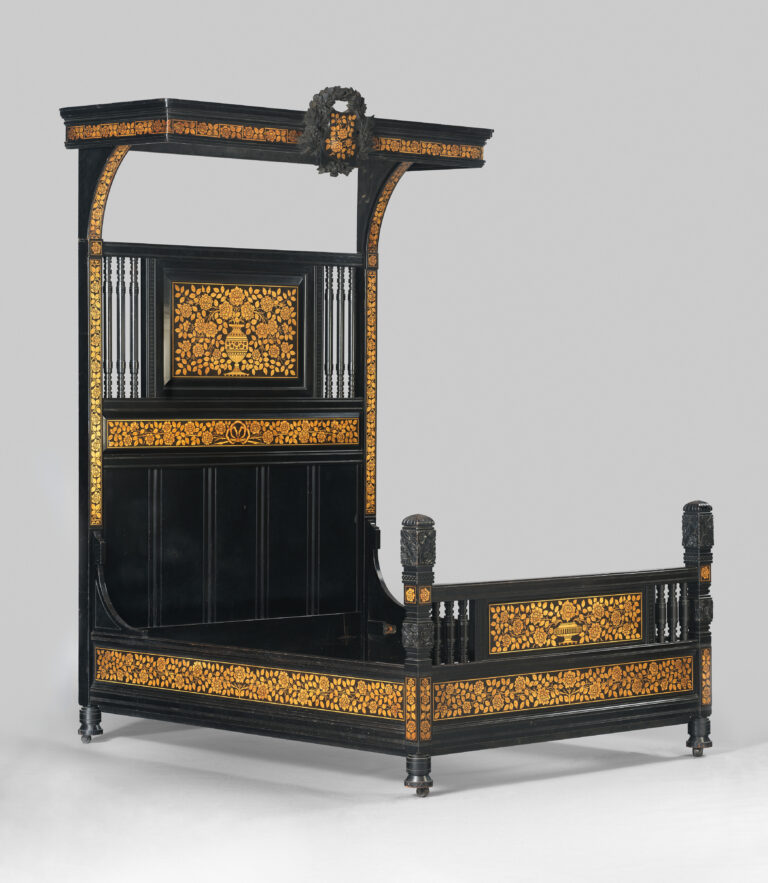 Victorian bedroom furniture: a tall ebonized bedstead with gilt ornament, a half canopy over the head and a panelled footboard, photographed against a grey studio ground