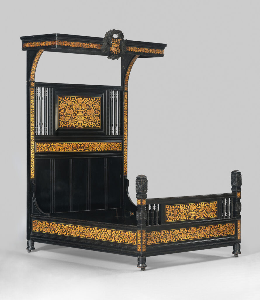 Victorian bedroom furniture: a tall ebonized bedstead with gilt ornament, a half canopy over the head and a panelled footboard, photographed against a grey studio ground