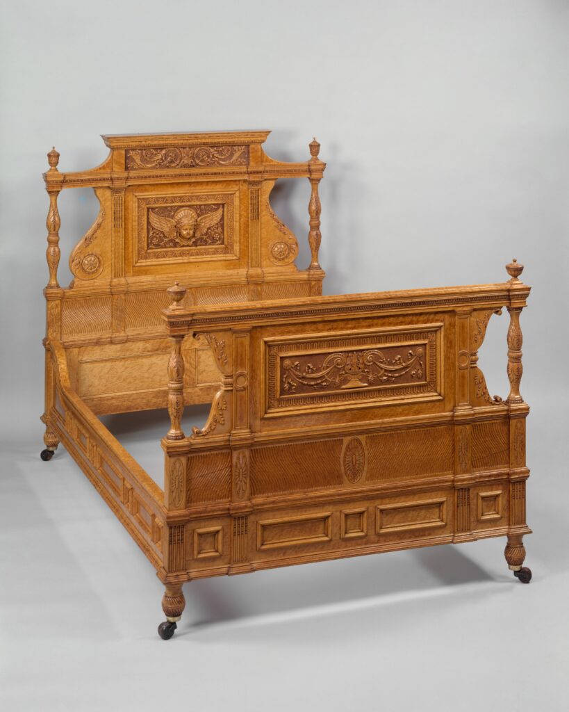 Light bird's-eye maple bedstead with a carved, panelled headboard and footboard, turned finials and small casters, against a neutral studio ground