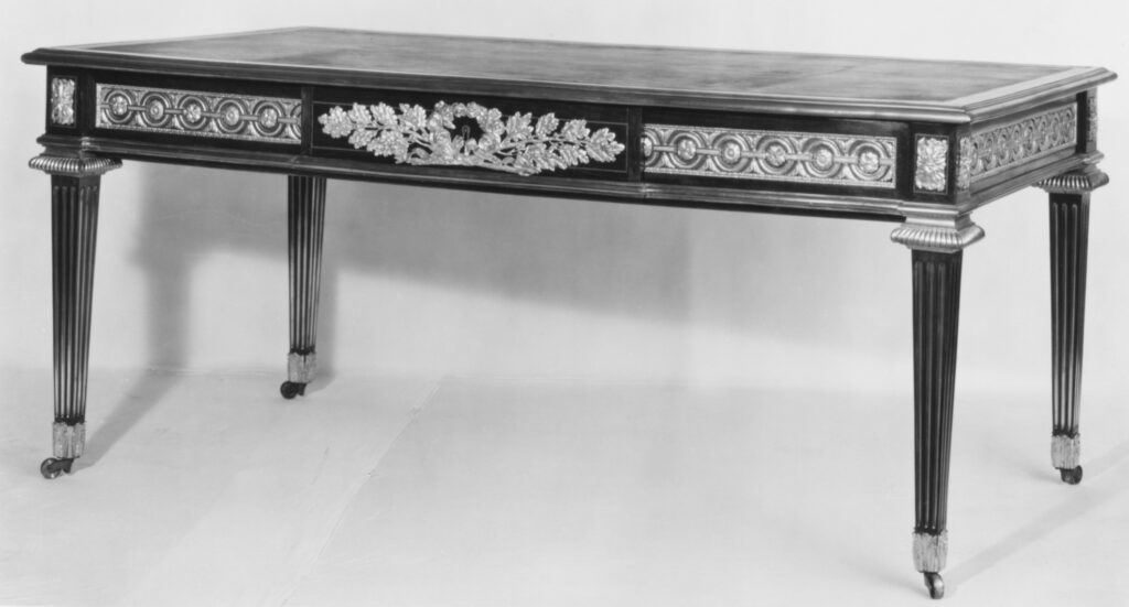 Black-and-white photograph of a long, plain writing table with a frieze of ornament, straight tapering fluted legs and a flat top