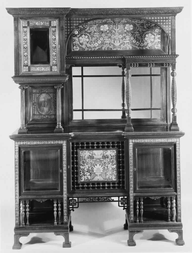 Black-and-white photograph of a tall display cabinet with turned spindles, glazed compartments of different sizes and an inlaid arched panel across the top