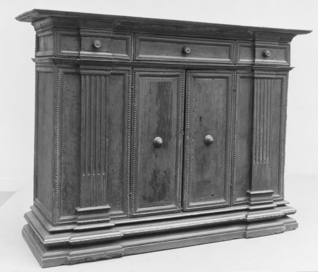 Black-and-white record photograph of a 16th-century walnut credenza: three drawers with round knobs under a projecting cornice, two panelled doors in the centre flanked by fluted pilasters, and a stepped plinth