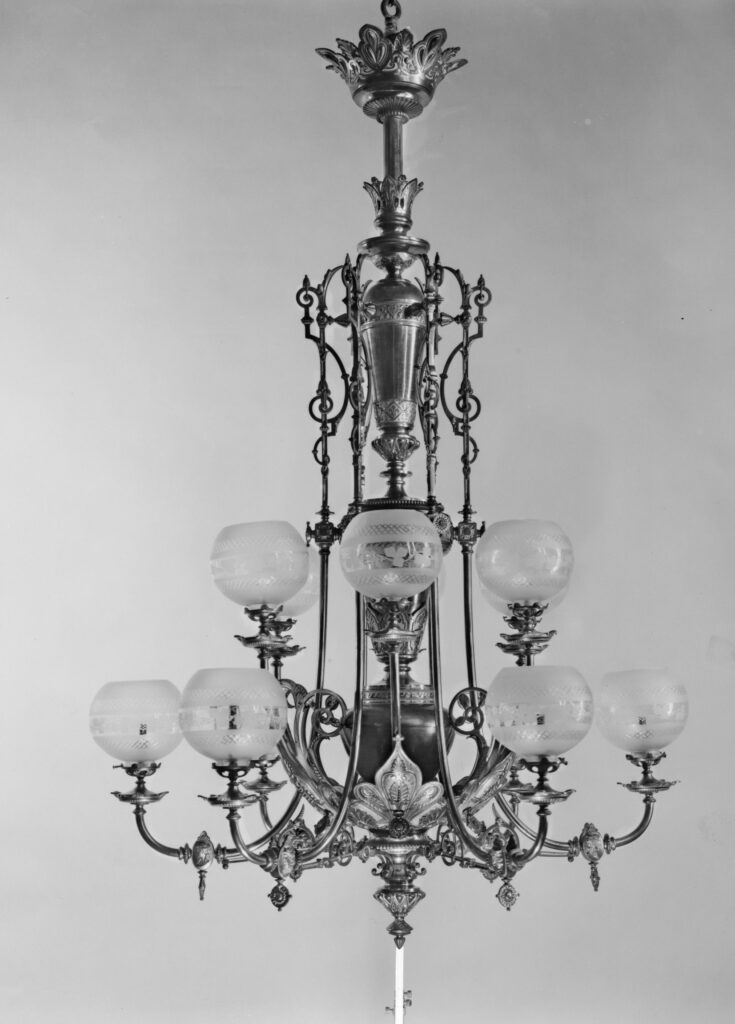 Black-and-white photograph of a tall brass chandelier with an open frame of scrolling metalwork and several round shades on curved arms