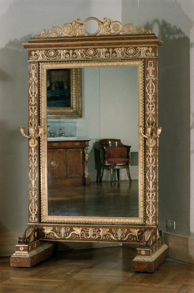 Empire style furniture: a tall cheval glass in an amboyna frame with gilt-bronze mounts, a crest with winged creatures and a frieze below the mirror, seen in a museum gallery