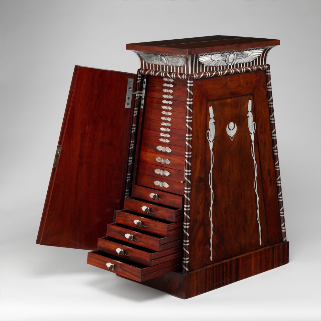 Tapering mahogany cabinet in the form of an Egyptian gateway, one door open to show rows of small drawers with silver mounts and a silver winged disk below the cornice