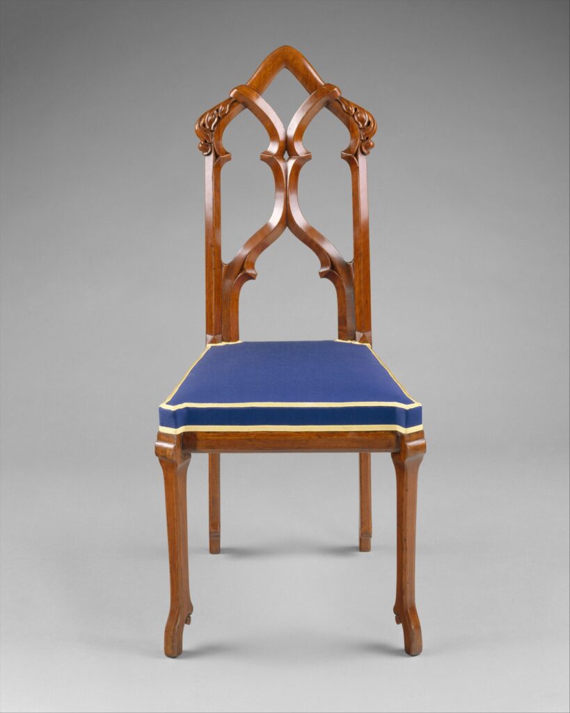 Wooden side chair with a back of two pointed arches that cross and curl like window tracery, carved ends at the top, a blue upholstered seat and slender curved front legs, on a gray ground