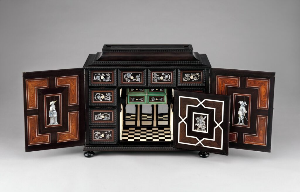 Small black ebony cabinet of about 1640-50 with both doors open: inside, rows of drawers faced with mother-of-pearl and tropical veneers surround a central compartment with a checkered floor and a mirrored back, the inner faces of the doors inlaid with small standing figures