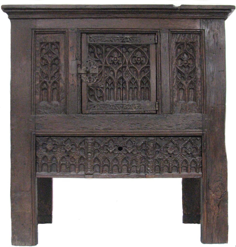 Dark oak French cupboard of about 1460 on tall end supports, its small central door and side panels carved with Gothic tracery of pointed arches, above a long frieze carved in the same pattern