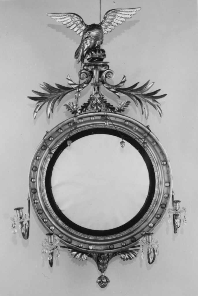 Black-and-white photograph of a round gilded mirror with a ball-studded frame, a spread-winged eagle at the top and candles standing in front of the glass