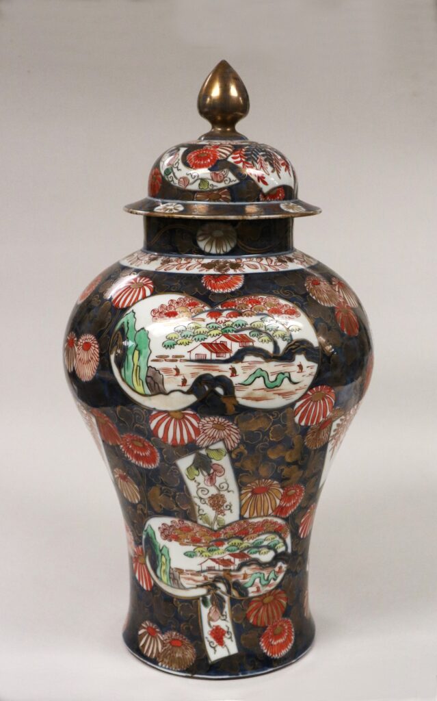 Tall Imari covered jar with a domed lid and gilt finial, painted on a dark ground with panels of flowers and scenes