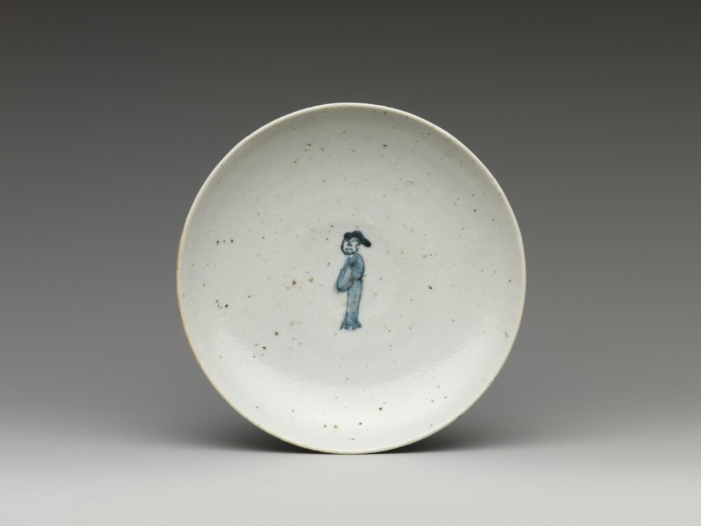 Shallow white dish with a small blue figure at the center and flecks in the glaze