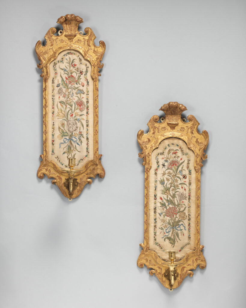Two tall narrow gilded wall sconces with carved shell-topped frames, each holding a panel of cream silk embroidered with flowers and a candle arm at the base