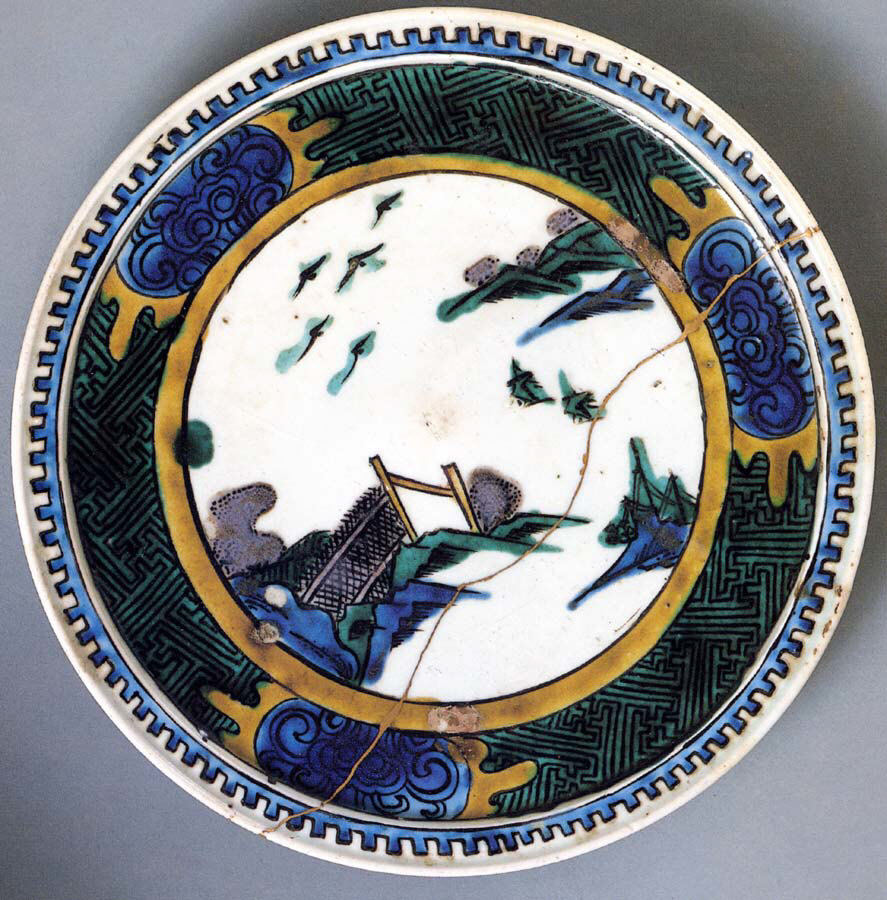 Round plate with a white center showing birds over rocks and a fence, inside a wide border of blue, green and gold patterns