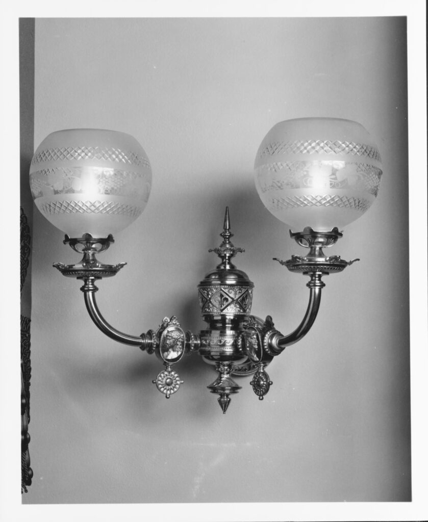 Black-and-white photograph of a brass wall bracket with two curved arms, each carrying a round frosted-glass globe with a cut band