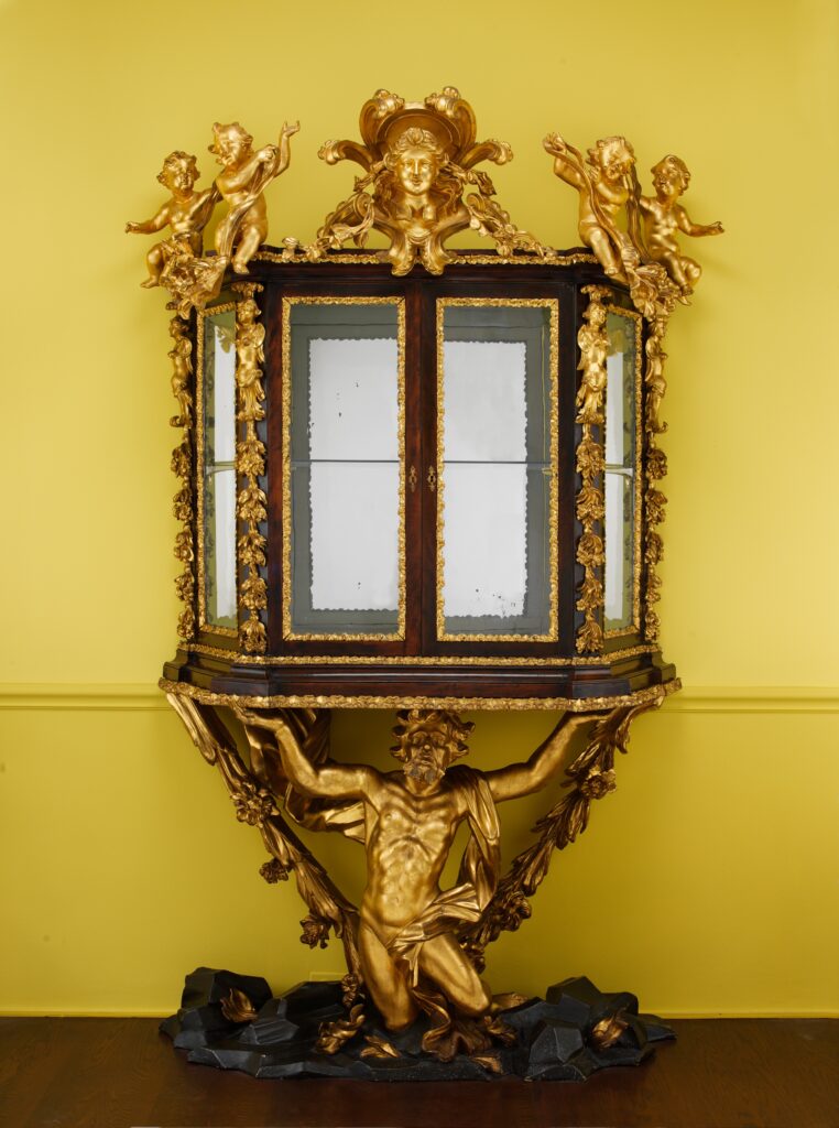 Roman Baroque furniture: a gilded and walnut display case with glazed doors, topped by small winged figures and a mask, carried on the shoulders of a kneeling gilded bearded figure on a dark green rocky base