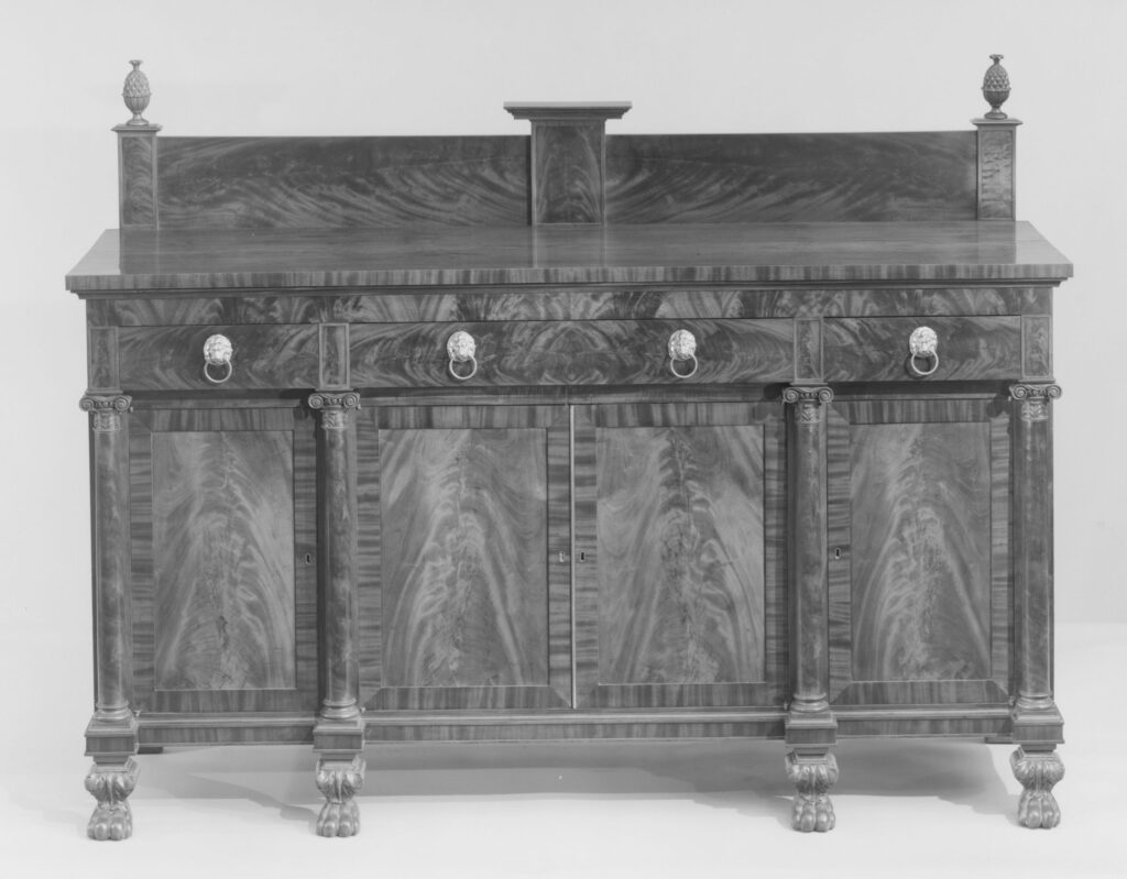 Black-and-white photograph of a heavy sideboard with a low back panel and two urn finials, a row of drawers above four tall veneered cupboard doors between columns, on paw feet