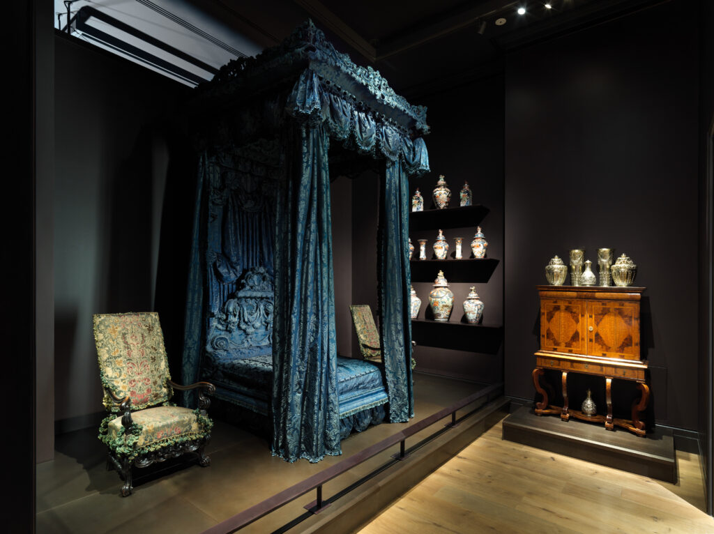 Baroque furniture: a tall state bed hung with blue silk damask, with a deep fringed canopy and curtains, shown in a dark museum gallery beside an upholstered armchair