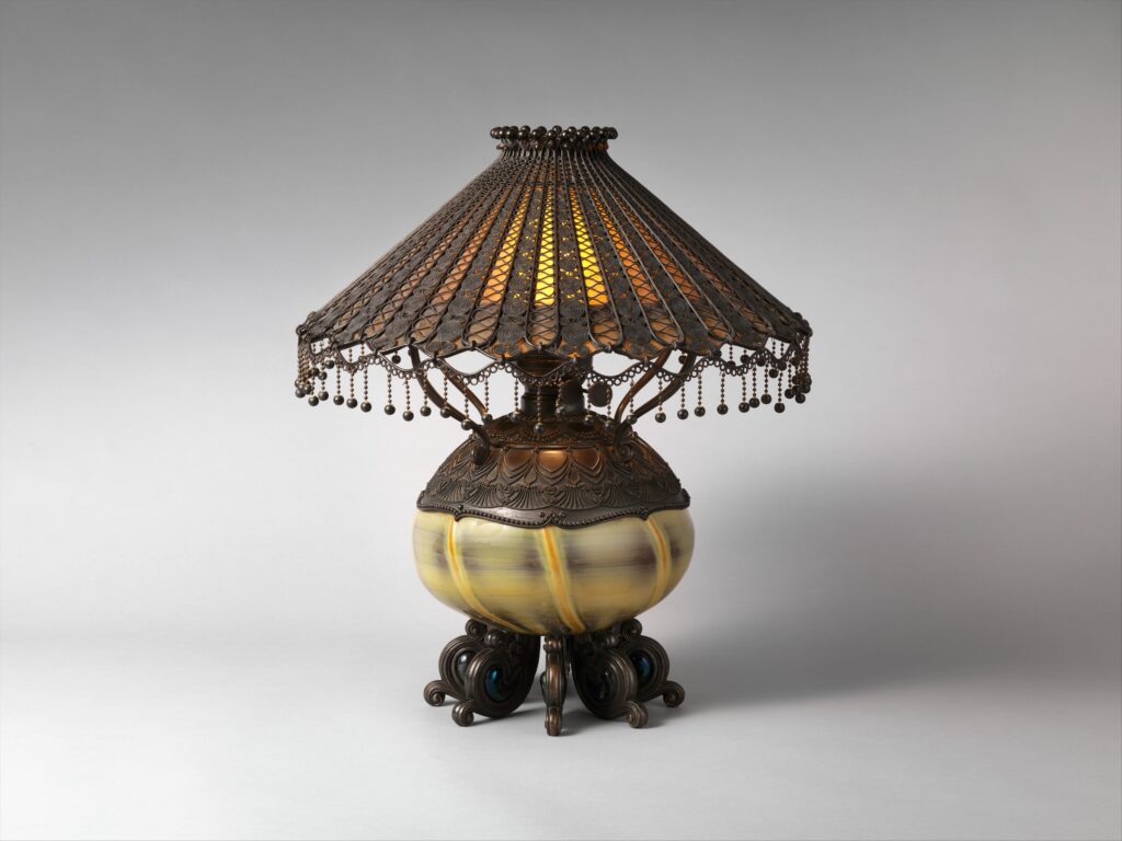 Table lamp with a conical shade of narrow bronze strips hung with small beads, over a ribbed pale yellow glass oil font on four scrolling bronze feet set with blue jewels