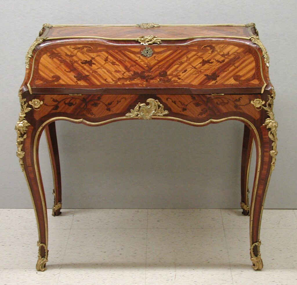 Small French slant-top desk of about 1745-49 on curving legs, its sloping lid veneered in a pattern of light and dark wood and edged with gilt bronze, with an open space beneath for the writer’s knees