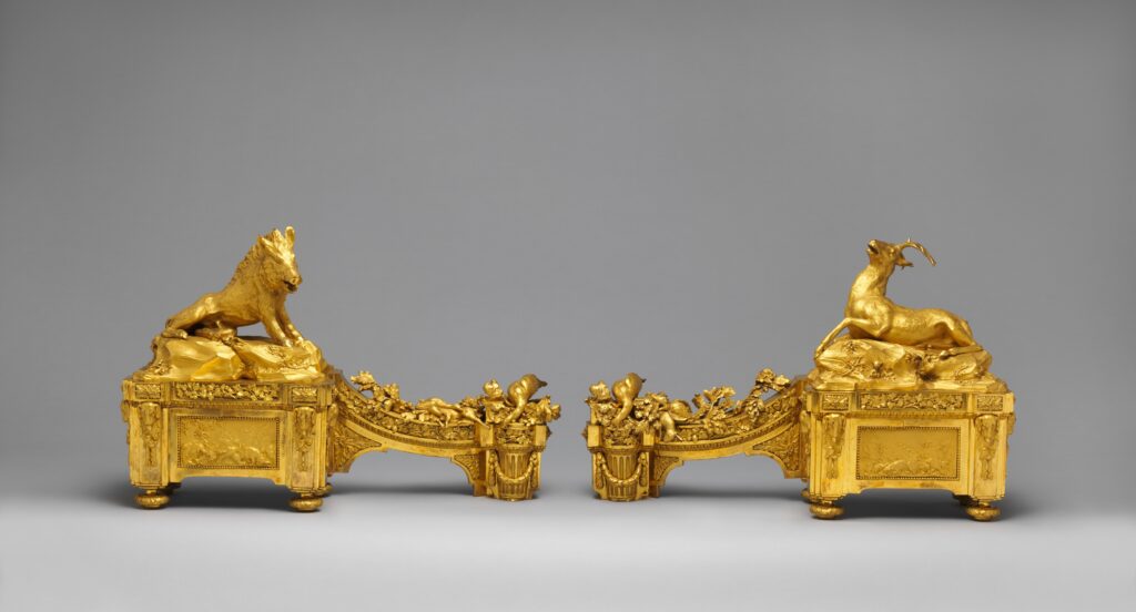 A pair of gilded firedogs, one topped by a boar and the other by a reclining stag, each on a rectangular base with a relief panel and joined by a bar of foliage and game, on a grey ground