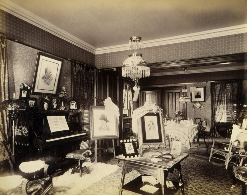 Sitting room photographs from 1888: a Victorian living room with an upright piano, framed portraits on easels, patterned wallpaper, a hanging glass-shaded lamp and a laid table in the next room